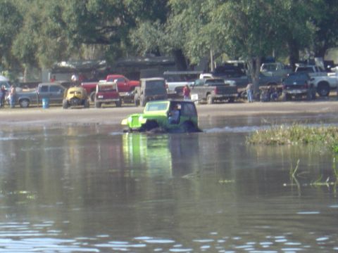 1987 Suzuki Samurai - Multiple off-road vehicles are gathered near a muddy body of water, with one green Jeep Wrangler actively navigating the water.