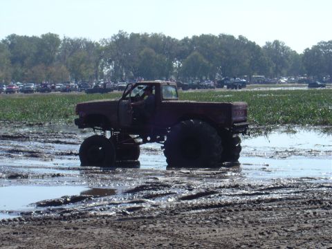 1987 Suzuki Samurai - Monster truck navigates a muddy terrain during an event, with spectators and parked vehicles visible.