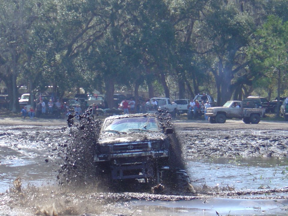 Truck driving through mud pit, splashing water. Other trucks and people in background