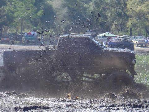 1987 Suzuki Samurai - A truck is engulfed in mud during an off-road event, with spectators and other vehicles visible in the distance.