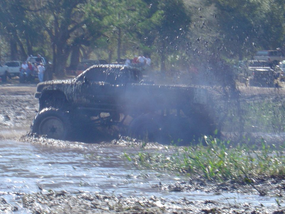 Muddy pickup truck driving through a mud pit, with other vehicles in the background