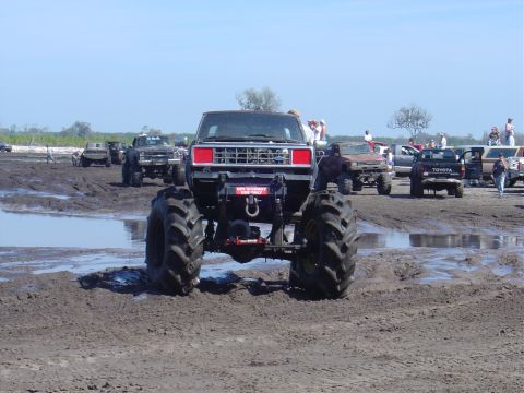 1987 Suzuki Samurai - Multiple modified trucks are seen in a muddy outdoor area under a clear blue sky.