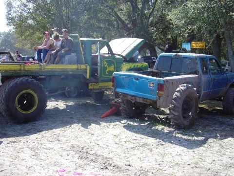 1987 Suzuki Samurai - Two heavily modified trucks with large tires are parked on sandy ground, with people riding in the back of the flatbed.