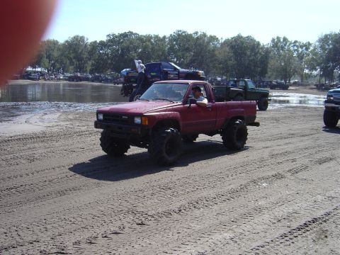 1987 Suzuki Samurai - Modified red pickup truck with oversized tires navigates a muddy track with other vehicles present.