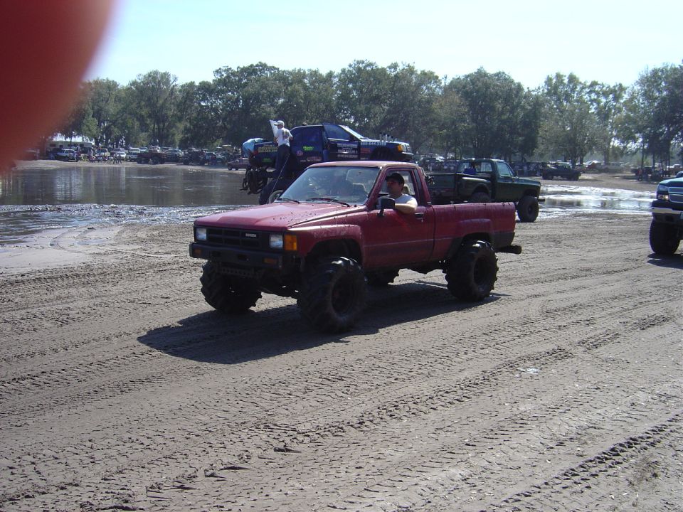 Red pickup truck with large tires driving on a dirt track. Other trucks are visible in the background