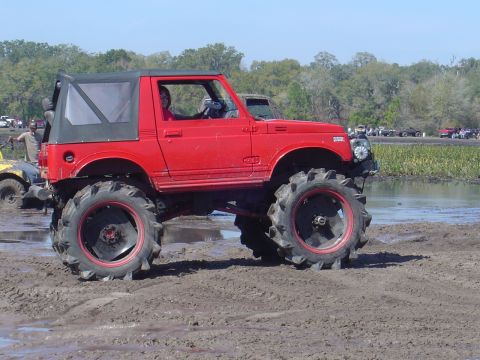 1987 Suzuki Samurai - A modified red 1987 Suzuki Samurai navigates a muddy terrain, with other vehicles visible in the background.