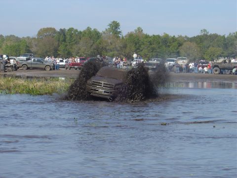 1987 Suzuki Samurai - A Dodge Ram truck navigates a mud bog, sending large plumes of mud into the air as onlookers watch from the shore.
