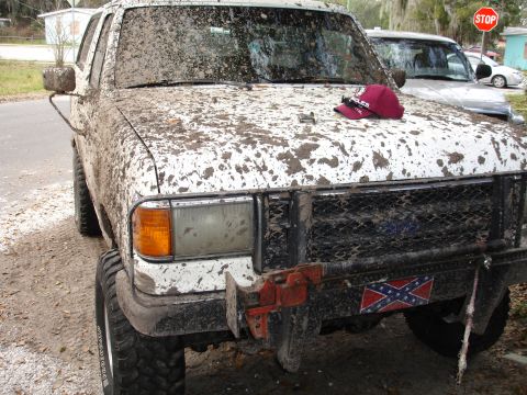 1989 Ford Bronco - A white SUV is heavily covered in mud, with a baseball cap resting on its hood.