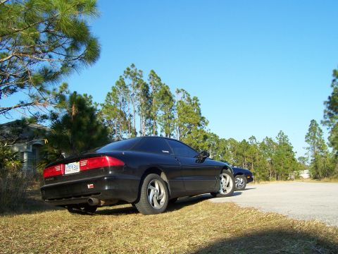 1994 Ford Probe - A black coupe is parked on a grassy area with trees in the background and another car behind it.