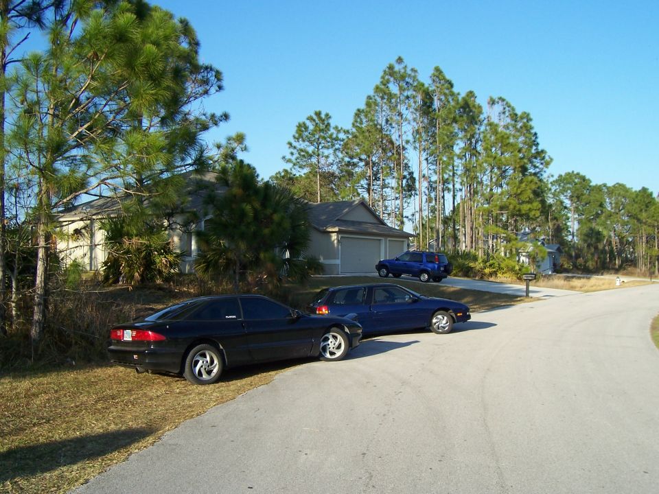 Black coupe parked on grass, blue sedan parked on road, house and trees in background