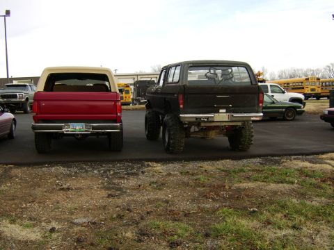 1978 Ford Bronco - Two SUVs are parked in a lot with other vehicles and school buses in the background.