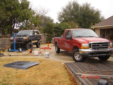 1991 Ford Bronco - A black Ford Bronco is being worked on with an engine hoist, while a red Ford pickup truck sits in the driveway.