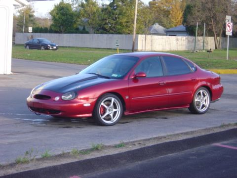 1998 Ford Taurus - A red sedan with a contrasting black hood is parked on a paved surface, with another vehicle visible in the distance.