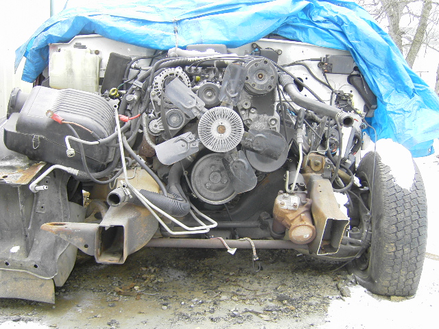 Front end of a truck chassis with engine exposed, covered partly by a blue tarp. Flat tire visible