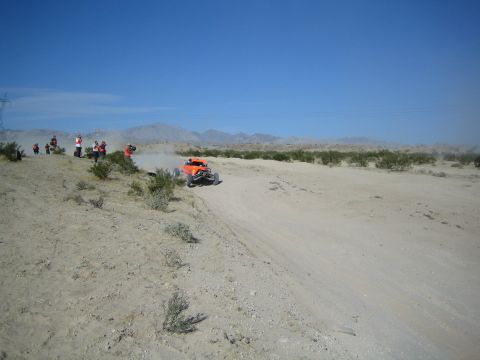 Orange off-road buggy kicking up dust near spectators in the desert