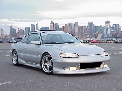 1994 Ford Probe - Silver coupe with body kit and spoiler parked in lot with city skyline.