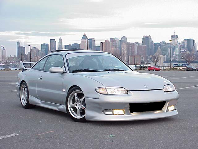 Silver coupe with aftermarket body kit and spoiler, parked in lot with city skyline behind