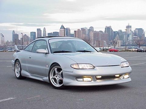 1994 Ford Probe - A silver Mazda MX-6 is parked in a lot with a city skyline visible behind it.