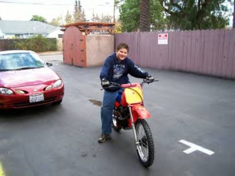 1972 Ford Ranchero - A red sedan is parked on asphalt next to a boy on a dirt bike.
