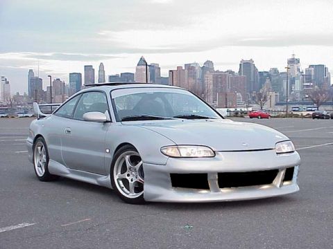 1994 Ford Probe - This silver coupe features an aggressive body kit and a rear spoiler, parked in a lot with a city skyline behind it.
