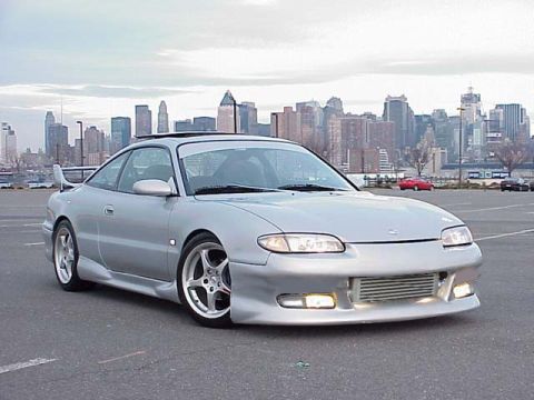 1994 Ford Probe - This silver coupe features an aggressive aftermarket body kit and a prominent rear spoiler, parked in a lot with a city skyline in the back…