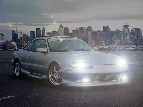 1994 Ford Probe - Silver coupe with aftermarket body kit and spoiler parked in front of a city skyline. Supermotors.net logo is visible.