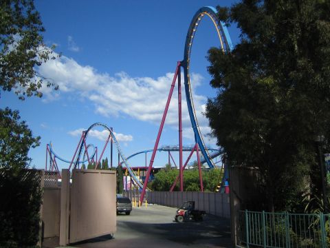 1990 Ford Bronco - blue-and-red looping steel roller coaster towers above trees and a service path under a partly cloudy sky.
