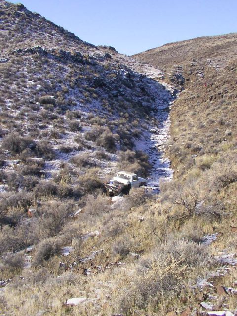 1978 Ford Bronco - A white pickup truck is stuck in a dry creek bed, surrounded by sparse vegetation and patches of snow on the surrounding hills.