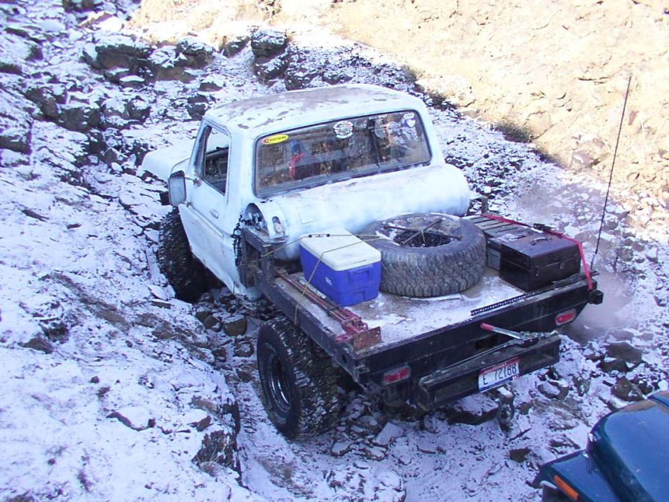 White flatbed truck with spare tire and cooler in the bed, stuck on a snowy, rocky incline