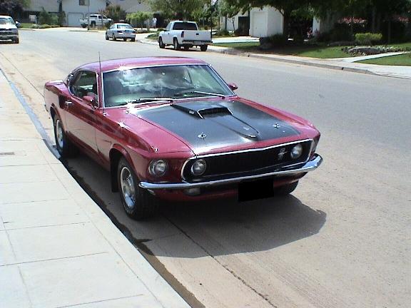 Front three-quarter of a red Mustang fastback with black hood and quad headlights at a curb