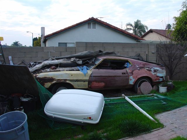 Rusty 1971 Ford Mustang fastback project car in primer, parked in a backyard under a tarp