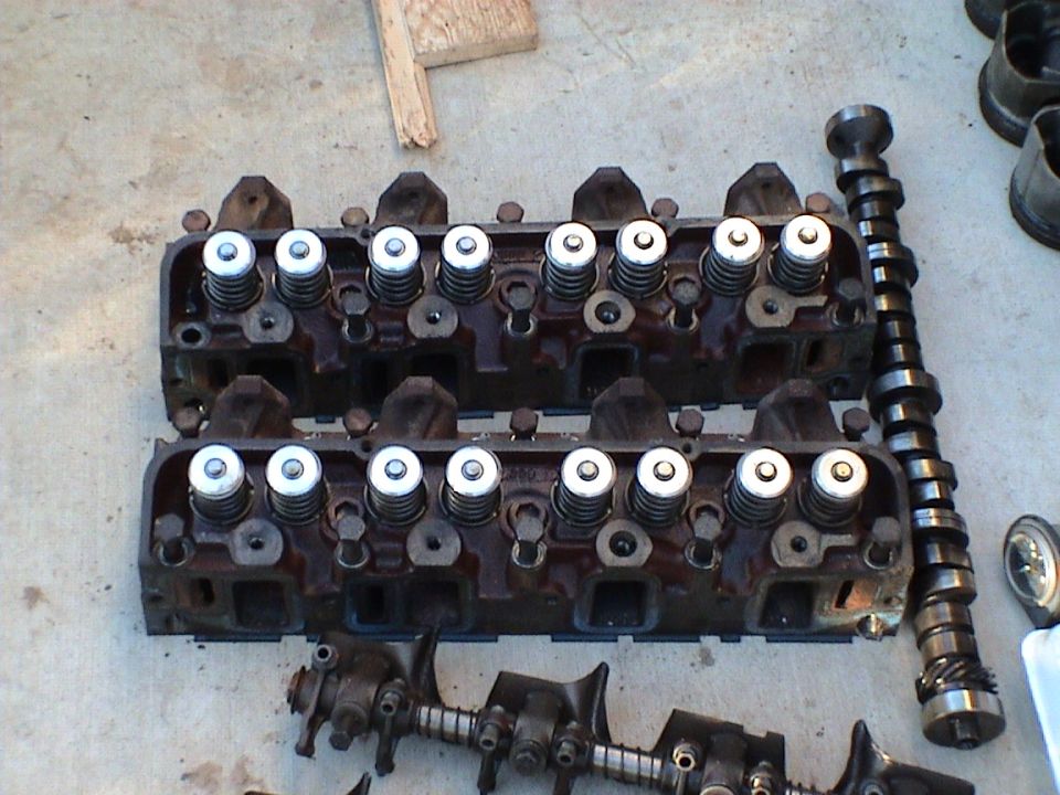 Two cast-iron cylinder heads with valves and springs installed lying beside a camshaft on concrete
