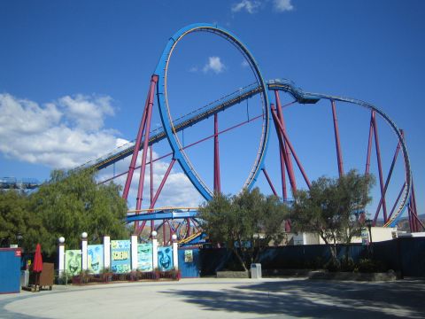 1990 Ford Bronco - blue-and-red steel roller coaster with a large vertical loop towers over a paved amusement park plaza under a clear sky.