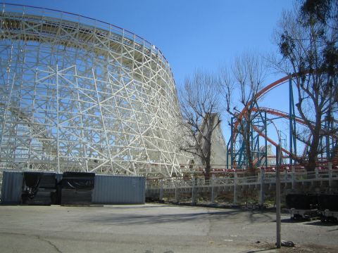1990 Ford Bronco - large white wooden roller coaster framework stands beside a red steel coaster track in a fenced amusement park lot.