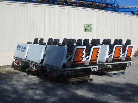 1990 Ford Bronco - Two roller coaster trains with orange-and-white cars and black restraint seats sit parked outside a building marked Training Trailer.
