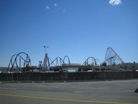 1990 Ford Bronco - Several roller coaster track silhouettes rise across an amusement park skyline beyond a fenced empty parking area.