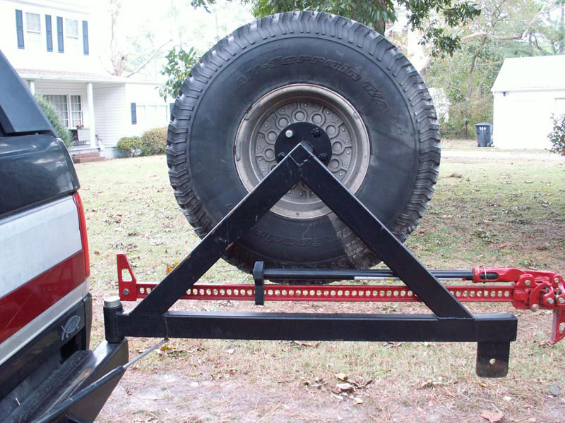 Black metal spare tire carrier with hi-lift jack attached to a red bracket, mounted on the rear of a red and white truck