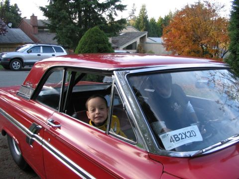1964 Ford Fairlane - A young boy smiles from the passenger seat of a red 1964 Ford Fairlane coupe, with another vehicle visible in the background.
