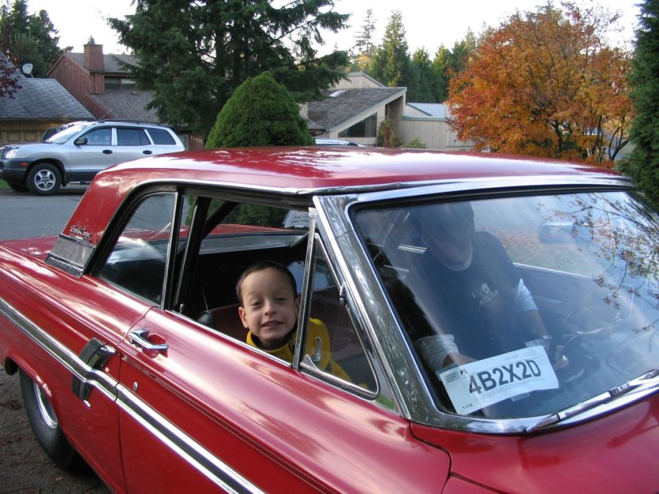 Boy leaning out window of red 1960s Ford two-door sedan in driveway