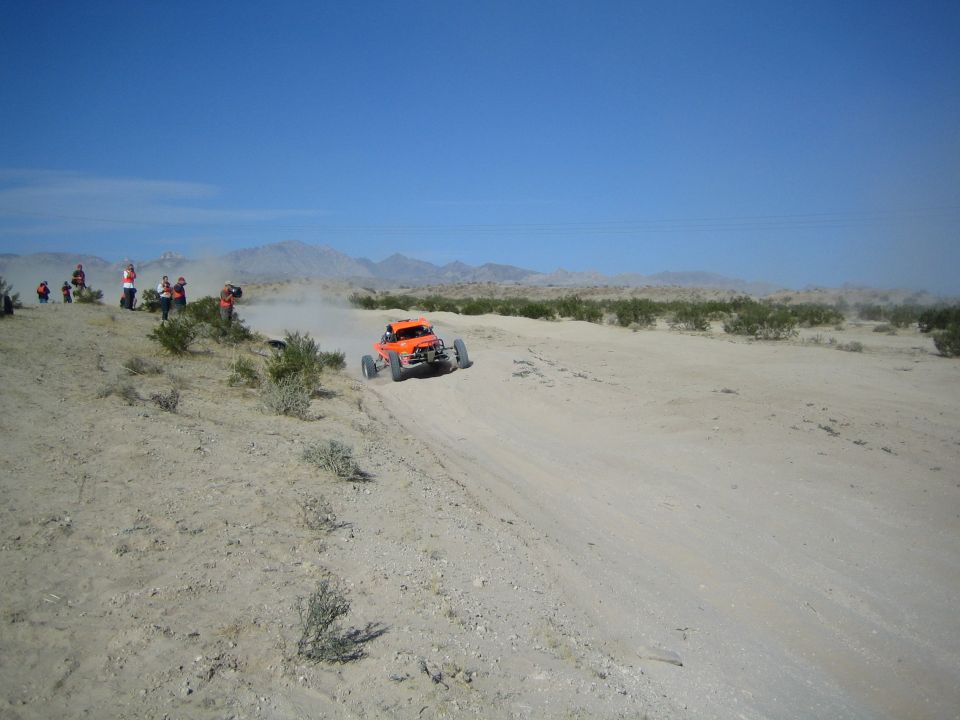 Orange off-road race buggy kicking up dust on a desert dirt course with spectators