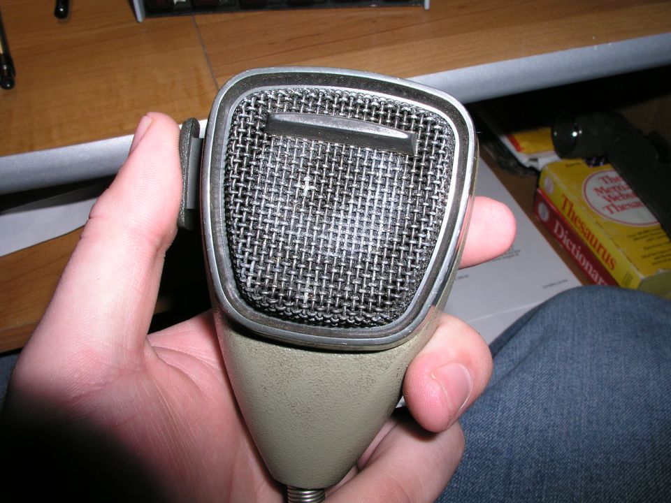 Hand holding a vintage microphone with a metal grille and beige handle. A dictionary is visible in the background
