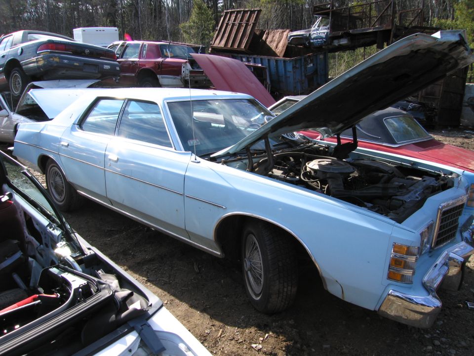 Light blue full-size sedan with hood up among junkyard cars