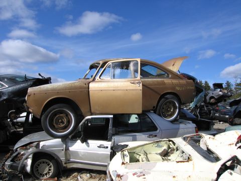 1991 Ford LTD Crown Victoria - tan fastback car perched on top of stacked wrecked vehicles in a junkyard under a blue sky.