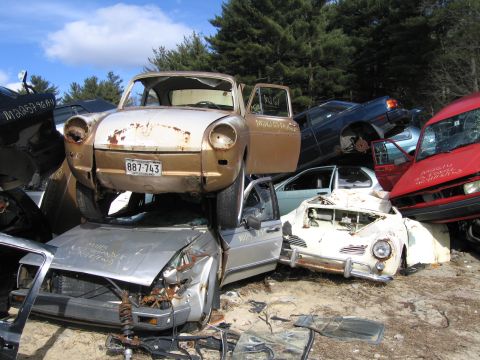 1991 Ford LTD Crown Victoria - rusted tan car body with a license plate reading 887-743 stacked atop other wrecked cars in a salvage yard.