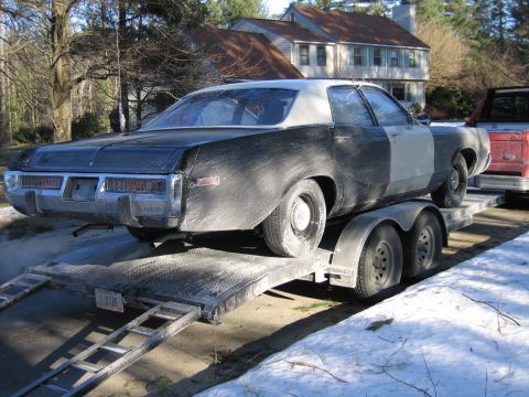 1991 Ford LTD Crown Victoria - dust-covered black two-door sedan with bare-metal primer patches sits loaded on a flatbed car trailer in a snowy suburban driveway.