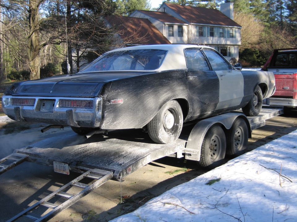 Rear of dusty black two-door sedan with primered panels on a tandem-axle car trailer in a driveway