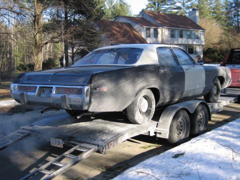 1991 Ford LTD Crown Victoria - Rear three-quarter view of a grimy black two-door coupe with primer patches secured on a tandem-axle flatbed trailer beside snow.