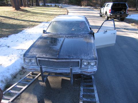 1991 Ford LTD Crown Victoria - Front view of a grimy dark full-size sedan with a chrome grille on ramps, its driver door open showing a white interior, a pickup behind.