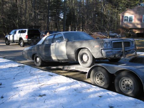 1991 Ford LTD Crown Victoria - dust-covered dark full-size sedan with a primered side stripe rests on a flatbed trailer hitched behind a white pickup near snow.