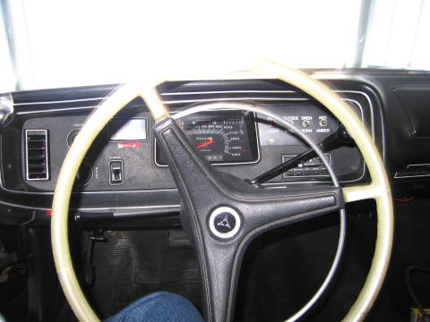 1991 Ford LTD Crown Victoria - Cream-colored steering wheel framing a dark dashboard with round speedometer and lettered warning indicators; a person's jeans are visible.